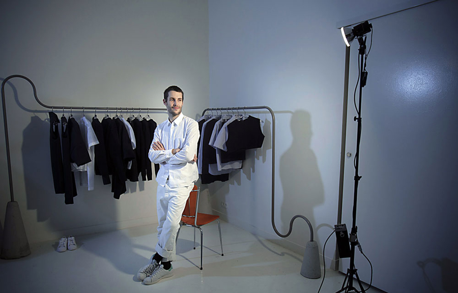 Simon Porte Jacquemus mặc đồ trắng, tạo dáng trong studio, sau lưng là rack treo quần áo.