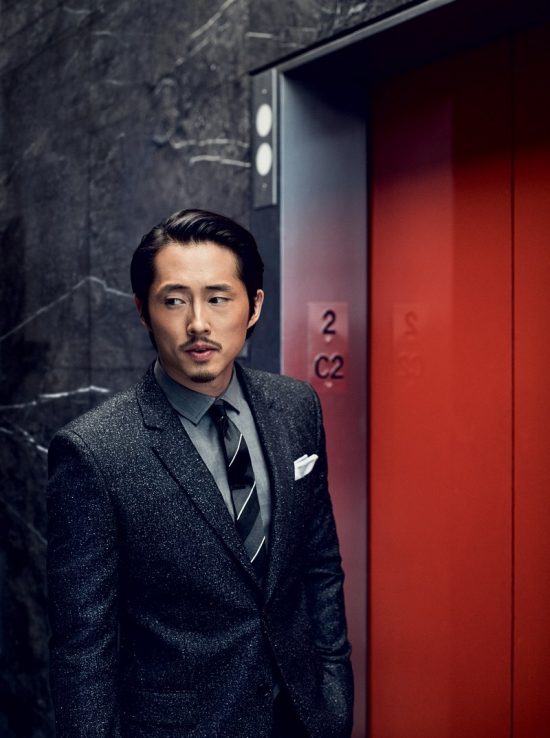 Steven Yeun trong bộ suit xám của Givenchy, đứng trước của thang máy màu đỏ.
