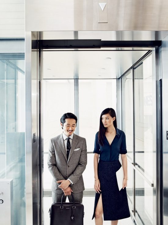Steven Yeun trong bộ tweed suit xám nhạt, đứng cùng cô gái áo xanh - váy xám xẻ tà.