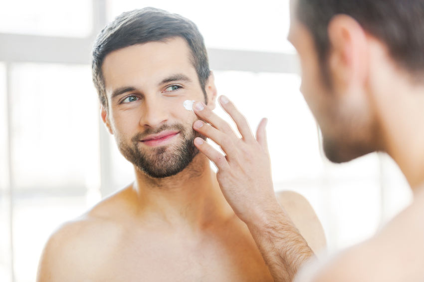 Skin care. Handsome young shirtless man applying cream at his face and looking at himself with smile while standing in front of the mirror